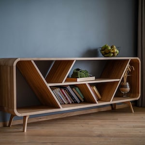 K&ouml;nnte beinhalten: Ein B&uuml;cherregal aus Holz mit einem einzigartigen geschwungenen Design und offenen Regalb&ouml;den. Das B&uuml;cherregal hat eine helle Holzoptik und weist ein geometrisches Muster auf den Regalb&ouml;den auf. Es steht auf einem Holzboden.