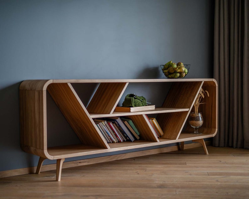 K&ouml;nnte beinhalten: Ein B&uuml;cherregal aus Holz mit einem einzigartigen geschwungenen Design und offenen Regalb&ouml;den. Das B&uuml;cherregal hat eine helle Holzoptik und weist ein geometrisches Muster auf den Regalb&ouml;den auf. Es steht auf einem Holzboden.