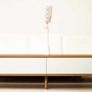 May include: A white couch with a wooden coffee table with two white drawers. A white vase with a dried white flower arrangement sits on the table.