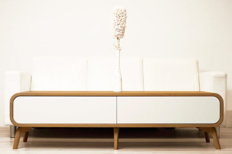 May include: A white couch with a wooden coffee table with two white drawers. A white vase with a dried white flower arrangement sits on the table.