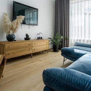 May include: A light blue sectional sofa with a wooden dresser and a television on the wall behind it. The sofa is in a living room with a light wood floor and a window with white curtains.