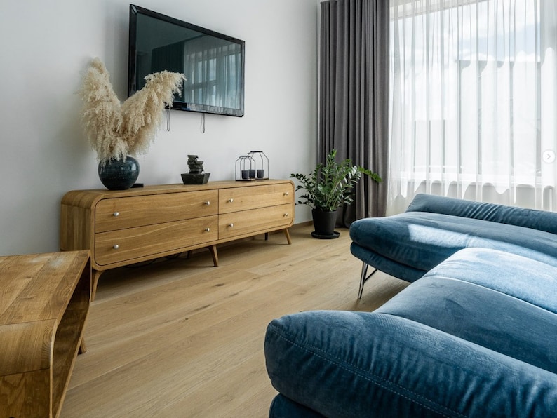 May include: A light blue sectional sofa with a wooden dresser and a television on the wall behind it. The sofa is in a living room with a light wood floor and a window with white curtains.