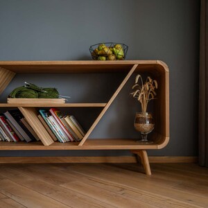 K&ouml;nnte beinhalten: Ein B&uuml;cherregal aus Holz mit geometrischem Design, mit drei Regalbrettern, die mit B&uuml;chern und einer gr&uuml;nen Wollkugel gef&uuml;llt sind. Das B&uuml;cherregal hat eine helle Holzoptik und steht auf vier Beinen. Eine Glasvase mit getrockneten Weizen&auml;hren steht auf dem oberen Regal.