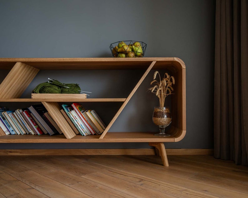 K&ouml;nnte beinhalten: Ein B&uuml;cherregal aus Holz mit geometrischem Design, mit drei Regalbrettern, die mit B&uuml;chern und einer gr&uuml;nen Wollkugel gef&uuml;llt sind. Das B&uuml;cherregal hat eine helle Holzoptik und steht auf vier Beinen. Eine Glasvase mit getrockneten Weizen&auml;hren steht auf dem oberen Regal.