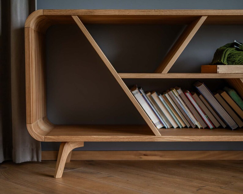 K&ouml;nnte beinhalten: Ein B&uuml;cherregal aus Holz mit einem einzigartigen geometrischen Design. Die Regale sind mit B&uuml;chern gef&uuml;llt und eine gr&uuml;ne Decke liegt auf dem obersten Regal.