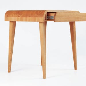 Puede incluir: Una mesa auxiliar de madera clara con una parte superior curvada y cuatro patas c&oacute;nicas. El caj&oacute;n est&aacute; parcialmente abierto, revelando su interior. La mesa est&aacute; hecha de madera natural y tiene un dise&ntilde;o minimalista.