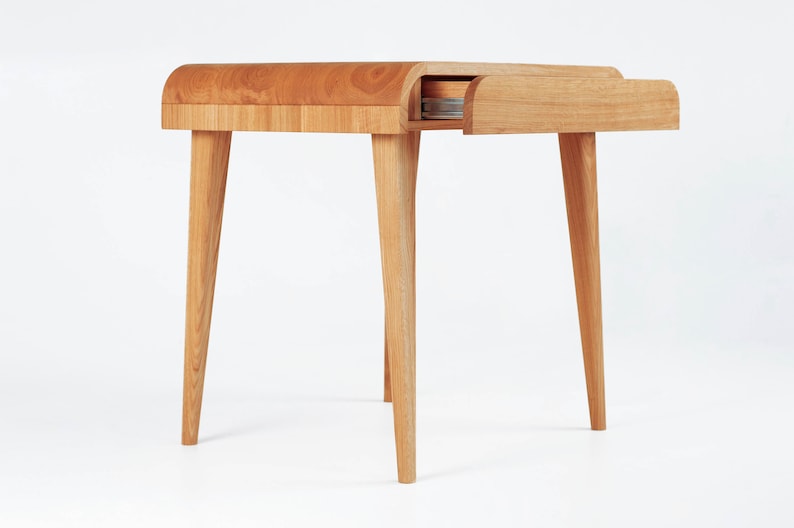 Puede incluir: Una mesa auxiliar de madera clara con una parte superior curvada y cuatro patas c&oacute;nicas. El caj&oacute;n est&aacute; parcialmente abierto, revelando su interior. La mesa est&aacute; hecha de madera natural y tiene un dise&ntilde;o minimalista.