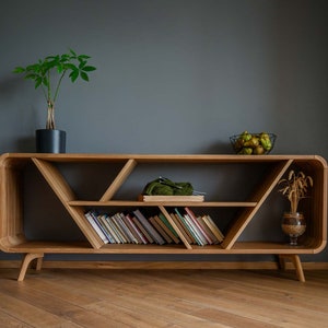 K&ouml;nnte beinhalten: Ein Holzregal mit geometrischem Design. Das Regal hat drei Ebenen und ist aus hellem braunem Holz gefertigt. In der untersten Ebene befinden sich B&uuml;cher, in der mittleren Ebene ein gr&uuml;nes Strickst&uuml;ck und in der obersten Ebene eine Schale mit Obst.