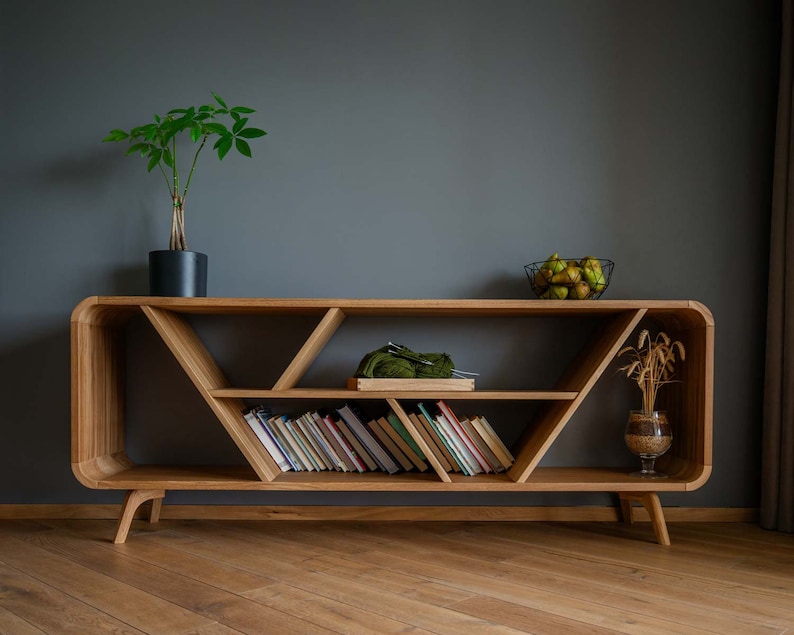 K&ouml;nnte beinhalten: Ein Holzregal mit geometrischem Design. Das Regal hat drei Ebenen und ist aus hellem braunem Holz gefertigt. In der untersten Ebene befinden sich B&uuml;cher, in der mittleren Ebene ein gr&uuml;nes Strickst&uuml;ck und in der obersten Ebene eine Schale mit Obst.