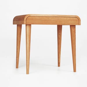 Puede incluir: Una mesa auxiliar de madera marr&oacute;n claro con una parte superior curvada y cuatro patas c&oacute;nicas. La mesa tiene un solo caj&oacute;n y est&aacute; hecha de madera maciza. El dise&ntilde;o es minimalista y moderno, adecuado para un sal&oacute;n o dormitorio.