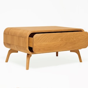 Peut inclure: Une table basse en bois clair avec un design incurvé et un seul tiroir. Le tiroir est ouvert et la table est posée sur un fond blanc.