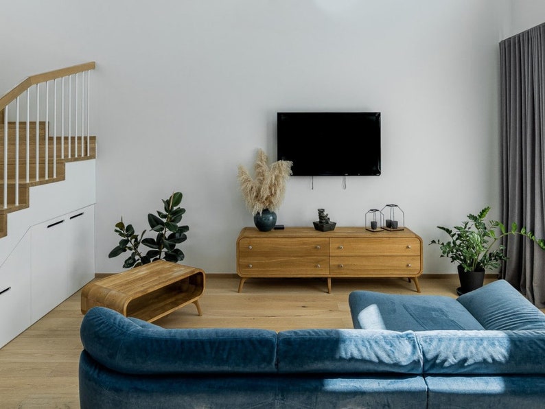 May include: A blue velvet sectional sofa with a wooden coffee table and a wooden dresser with a flat screen TV mounted above it. There are plants and a vase with dried flowers on top of the dresser.