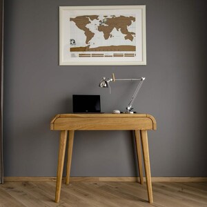 Puede incluir: Un escritorio de madera clara con un port&aacute;til, una l&aacute;mpara de escritorio plateada y un rat&oacute;n blanco. Un mapa del mundo enmarcado cuelga de la pared gris. El escritorio tiene cuatro patas anguladas y un borde frontal curvo. Mide unos 120 cm de largo.