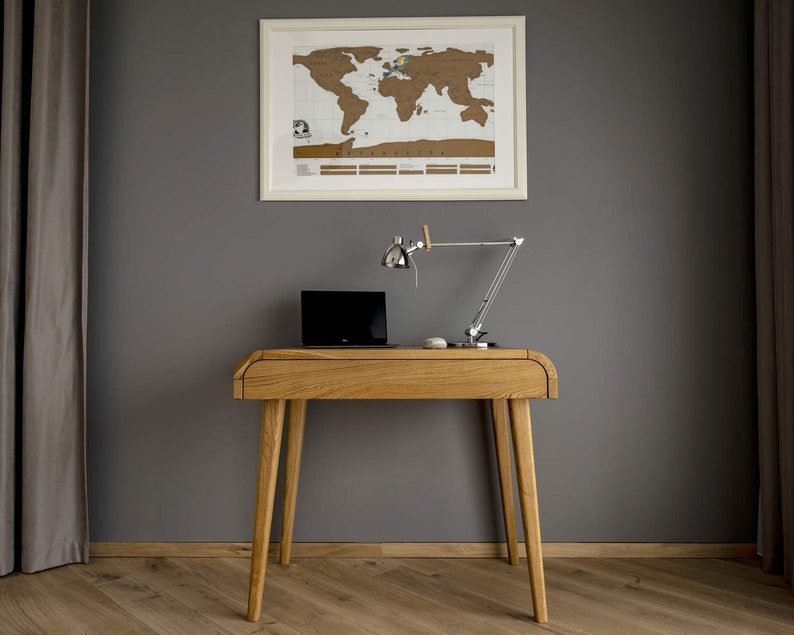 Puede incluir: Un escritorio de madera clara con un port&aacute;til, una l&aacute;mpara de escritorio plateada y un rat&oacute;n blanco. Un mapa del mundo enmarcado cuelga de la pared gris. El escritorio tiene cuatro patas anguladas y un borde frontal curvo. Mide unos 120 cm de largo.