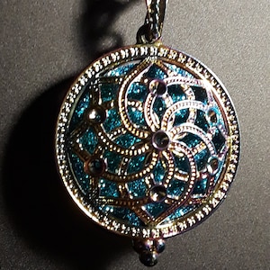 Puede incluir: Un colgante redondo de color dorado con un diseño de filigrana azul verdoso y plateado. El colgante tiene un patrón detallado e intrincado y está unido a una cadena.