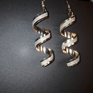 Puede incluir: Un par de pendientes de color dorado con un diseño en espiral. Los pendientes están adornados con purpurina plateada.