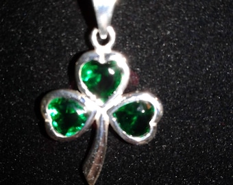Puede incluir: Un collar de plata con un colgante de trébol verde. El trébol tiene tres hojas, cada una con una piedra preciosa verde.