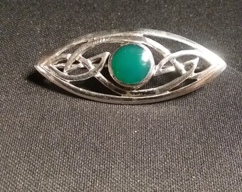 Pode incluir: Um broche de prata com um nó celta e uma pedra verde no centro.