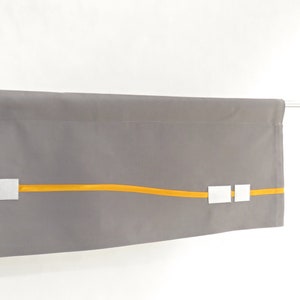 Peut inclure: Une cantonnière en tissu gris avec un ruban jaune et des carrés argentés. La cantonnière est suspendue à une tringle blanche.