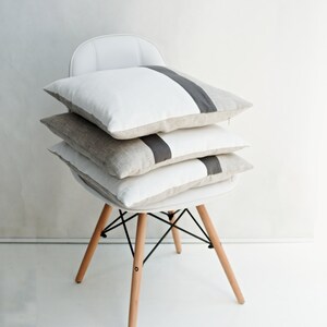 Puede incluir: Tres almohadas decorativas apiladas en una silla blanca. Las almohadas son de lino y tienen un diseño de rayas grises.