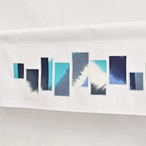 Peut inclure: Une cantonnière blanche avec un motif géométrique de rectangles bleus et turquoise. La cantonnière est suspendue à une tringle argentée.