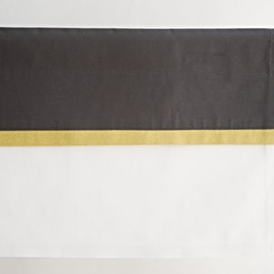 Peut inclure: Un cantonnière de fenêtre moderne avec un design en trois tons. La partie supérieure est gris foncé, suivie d'une bande dorée et d'un bas blanc. La cantonnière est suspendue à une tringle argentée, créant un look épuré et contemporain.