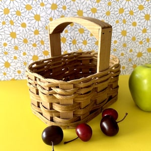 May include: A small, woven basket with a wooden handle. The basket is light brown and sits on a yellow surface, with green apples and cherries nearby. The background features a daisy pattern.
