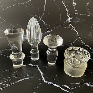 May include: Four clear glass bottle stoppers of varying designs. One is a small, flared cup shape. Another is a tall, ribbed teardrop. The third is a round, domed shape. The last is a circular stopper with a scalloped edge.