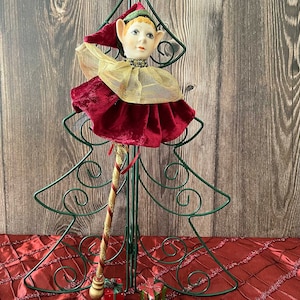 May include: A porcelain elf doll with a red and gold costume, holding a candy cane, is perched on a green metal Christmas tree stand.