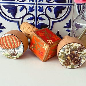 May include: Three decorative bottle stoppers with different designs. One features a hot air balloon, another has a floral pattern, and the third is a red and gold geometric design.
