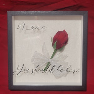 May include: A framed artwork featuring a red rose with a white ribbon, set against a textured off-white background. The frame is dark gray. The words "Name" and "You should be here" are written in cursive.