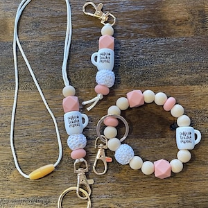 May include: A set of three teacher-themed keychains with a white coffee cup charm that says "coffee teach repeat". The keychains are made with wooden beads, pink and white silicone beads, and white crocheted beads. The keychains are attached to gold key rings.