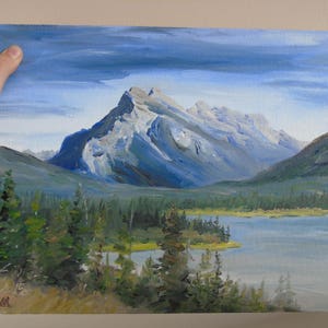May include: A painting of a mountain range with a lake in the foreground. The mountain peaks are covered in snow and the sky is a light blue with clouds. The lake is a light blue and there are trees in the foreground.
