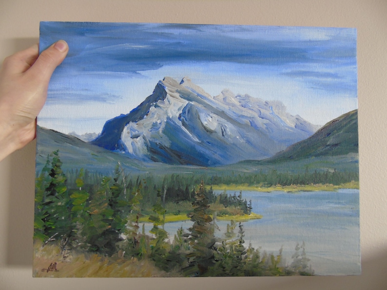 May include: A painting of a mountain range with a lake in the foreground. The mountain peaks are covered in snow and the sky is a light blue with clouds. The lake is a light blue and there are trees in the foreground.