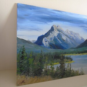 May include: A painting of a mountain range with a lake in the foreground. The mountains are a light blue and gray color, and the lake is a dark blue. The sky is a light blue with some clouds. There are trees in the foreground.