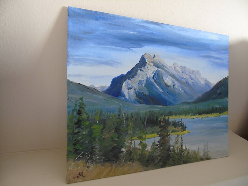 May include: A painting of a mountain range with a lake in the foreground. The mountains are a light blue and gray color, and the lake is a dark blue. The sky is a light blue with some clouds. There are trees in the foreground.