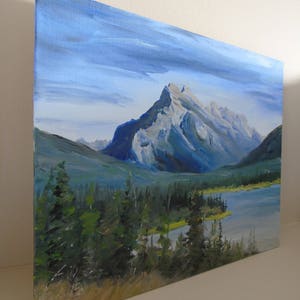 May include: A painting of a mountain range with a lake in the foreground. The mountains are a light gray color with blue and green hues. The lake is a dark blue color with a light green shoreline. The sky is a light blue color with white clouds.