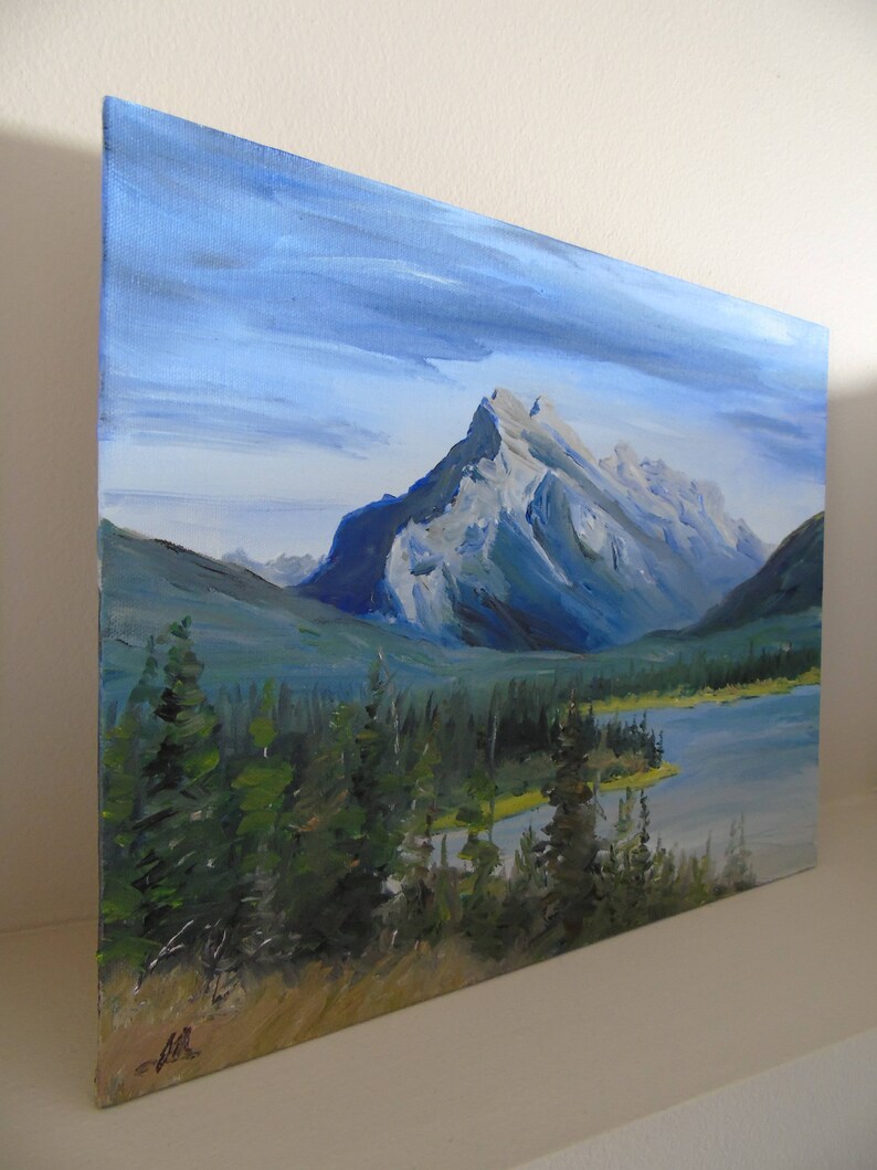 May include: A painting of a mountain range with a lake in the foreground. The mountains are a light gray color with blue and green hues. The lake is a dark blue color with a light green shoreline. The sky is a light blue color with white clouds.