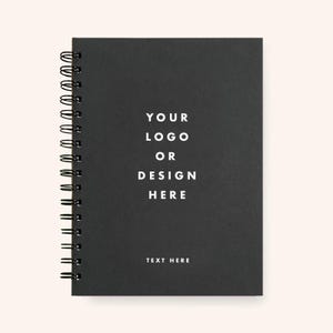 Puede incluir: Un cuaderno negro con espiral y el texto blanco "YOUR LOGO OR DESIGN HERE" y "TEXT HERE". El cuaderno tiene un acabado mate y encuadernación de alambre en el lado izquierdo. El fondo es de color beige claro.