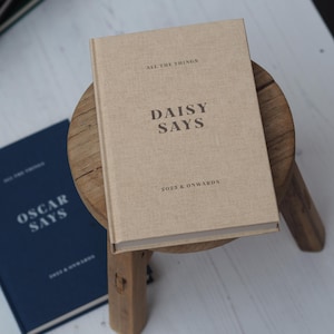 May include: A tan hardcover journal with the text "DAISY SAYS" and "2025 & ONWARDS" is on a small wooden stool. A navy blue journal with the text "OSCAR SAYS" and "2025 & ONWARDS" is next to it. The journals are for note-taking.