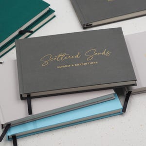 Può includere: Una pila di quaderni con copertina rigida in vari colori, tra cui grigio scuro, grigio chiaro, verde acqua e azzurro. Il quaderno grigio scuro in alto presenta la scritta in lamina d'oro Scattered Sands SAFARIS & EXPEDITIONS e un segnalibro nero.