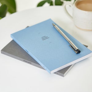 May include: A stack of notebooks with the top one in light blue, featuring the text "YOGA TEACHER TRAINING". A silver pen rests on the blue notebook. A gray notebook with the text "PODCASTS" is visible below. A mug of coffee sits in the background.