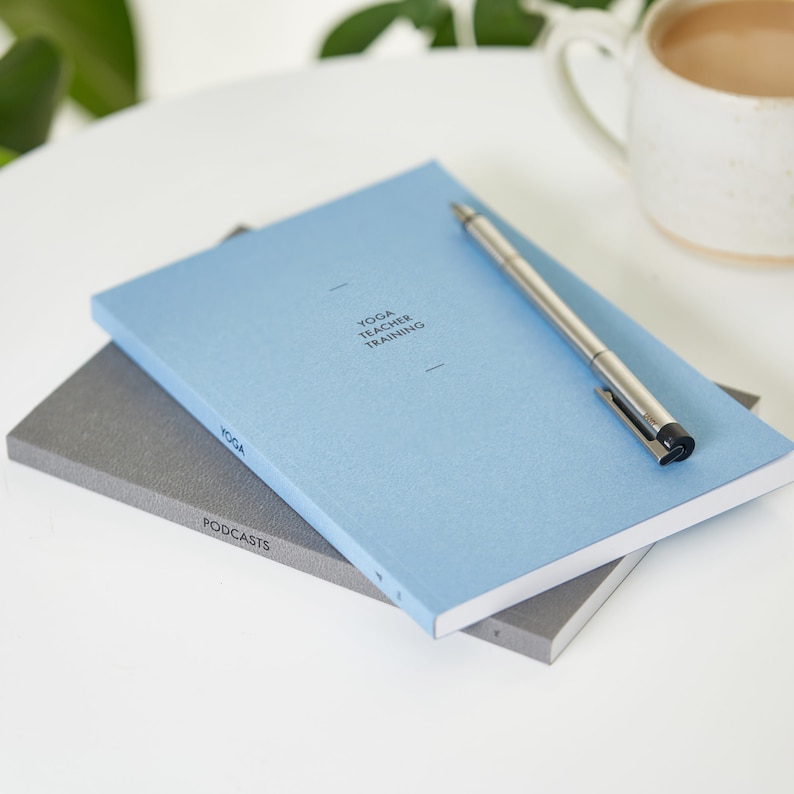 May include: A stack of notebooks with the top one in light blue, featuring the text "YOGA TEACHER TRAINING". A silver pen rests on the blue notebook. A gray notebook with the text "PODCASTS" is visible below. A mug of coffee sits in the background.