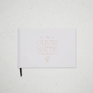Könnte beinhalten: Ein weißes Gästebuch im Querformat mit goldener Folienprägung, die OLIVER AND HATTIE, 05.06.2027, und WELL WISHES FROM FRIENDS & FAMILY zeigt. Unter dem Text befindet sich eine kleine goldene Blumenzeichnung, und ein schwarzes Lesebändchen hängt unten heraus.