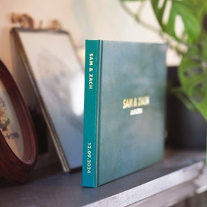 May include: A dark green hardcover photo album with gold foil lettering on the front and spine. The text on the front reads SAM & ZACH and 12.09.2024, while the spine features the text SAM & ZACH and 12.09.2024 in a vertical orientation.