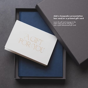 May include: A navy blue journal and a white card with gold lettering that reads "A GIFT FOR YOU" are presented inside a gray gift box. The box is partially open, revealing the contents. The text on the card suggests a gift-giving theme.