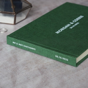 Puede incluir: Un libro de visitas de tapa dura verde con el texto "MR. & MRS. BARTHORPE" y "MORGAN & CORRIE 20.10.2023" impreso en blanco.