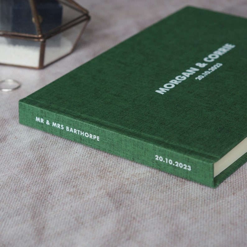 Puede incluir: Un libro de visitas de tapa dura verde con el texto "MR. & MRS. BARTHORPE" y "MORGAN & CORRIE 20.10.2023" impreso en blanco.