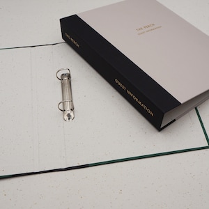 Op de afbeelding: Een gasteninformatie-map met een lichtgrijze kaft en een zwarte rug. De tekst "THE PERCH" en "GUEST INFORMATION" zijn in goud gedrukt. Een zilveren bindring is zichtbaar.