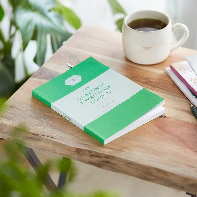 Pu&ograve; includere: Un diario verde con il titolo "My Drawings & Writings, Aged 2" e il nome "Noah Blay". Il diario &egrave; aperto su un tavolo di legno, accanto a una tazza di t&egrave; e altri libri.